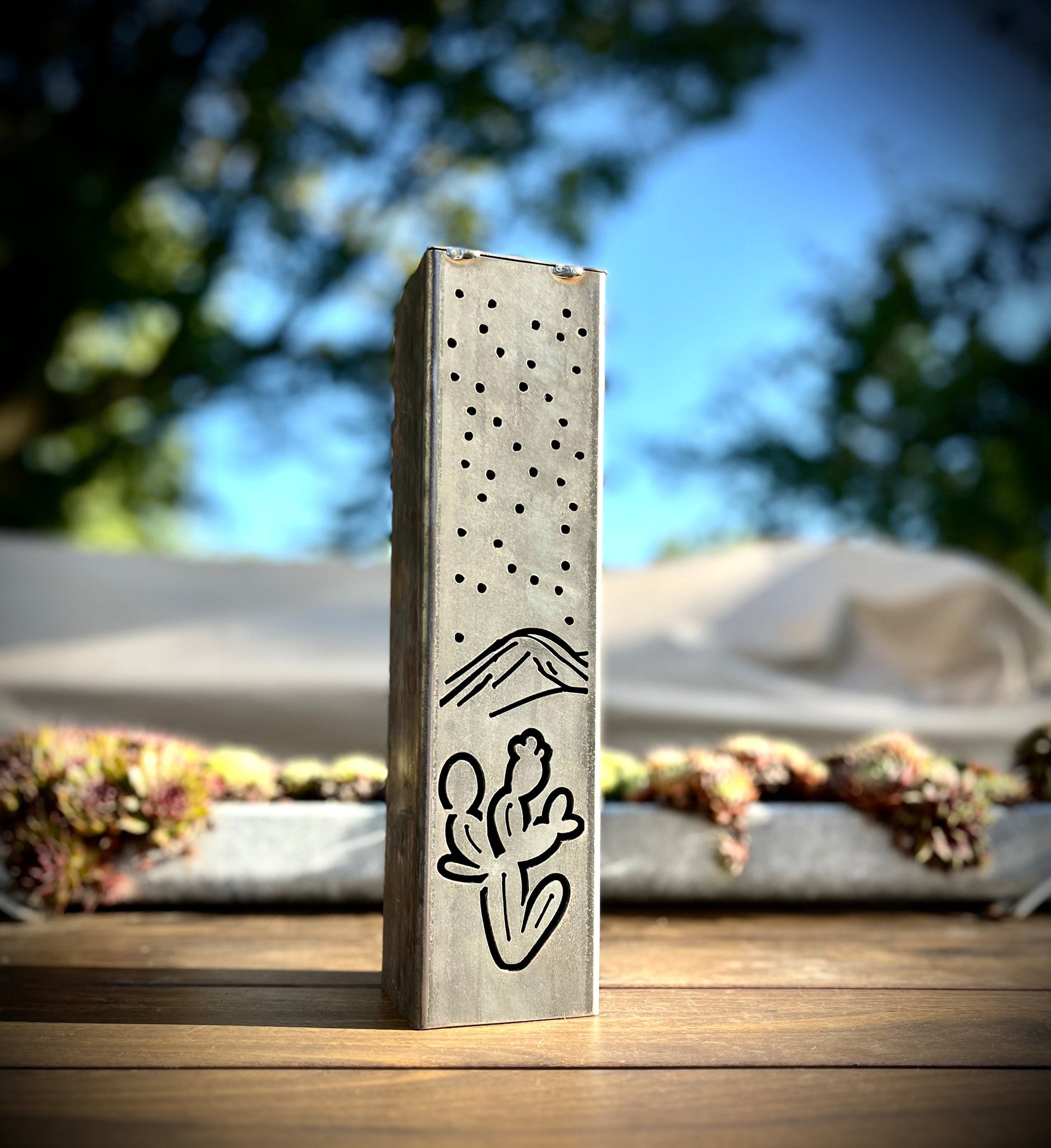Decorative metal pillar with cut-out patterns on a wooden surface with a blurred outdoor background.