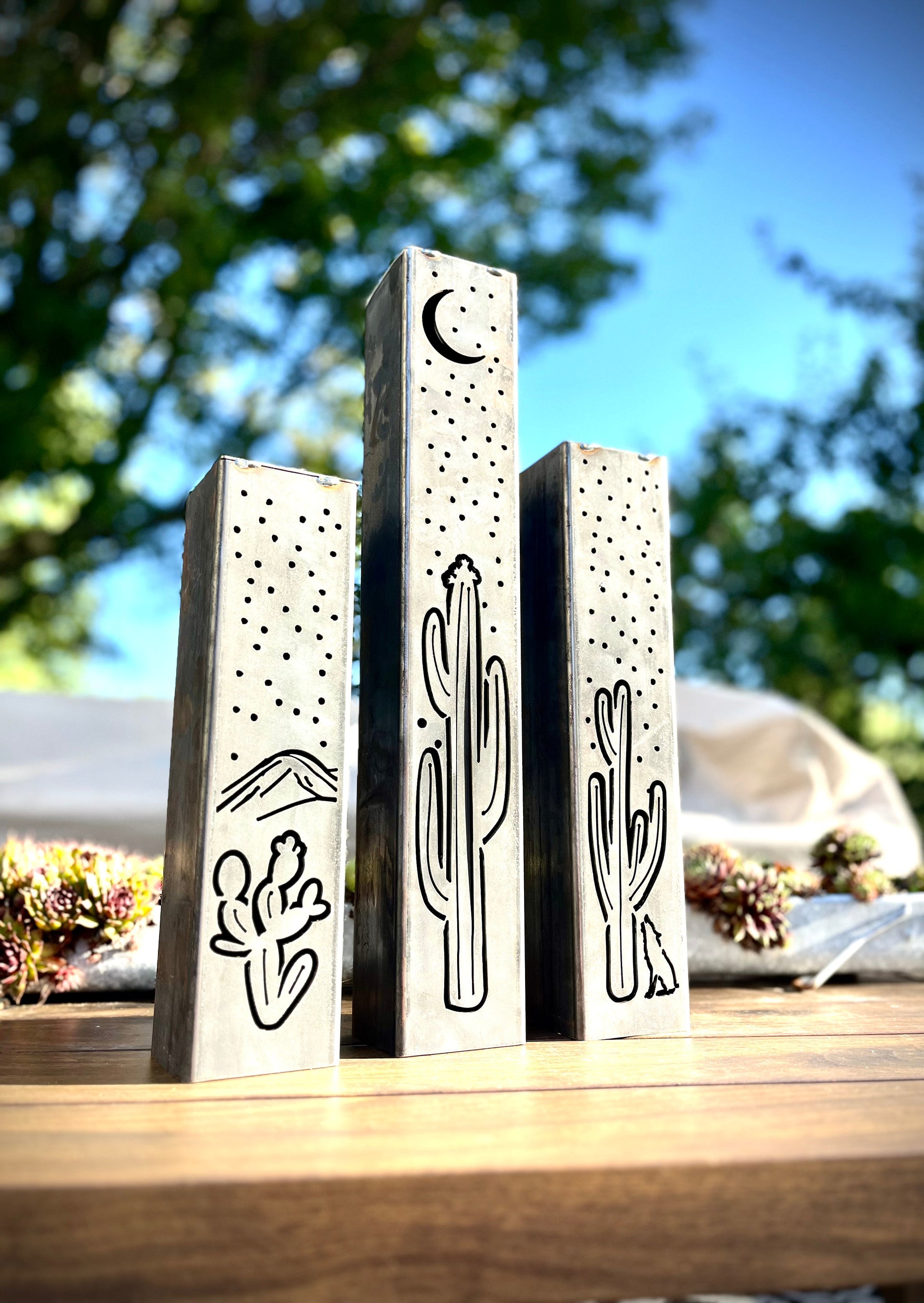 Three decorative metal pillars with cactus and moon designs on a wooden surface with a blurred natural background.
