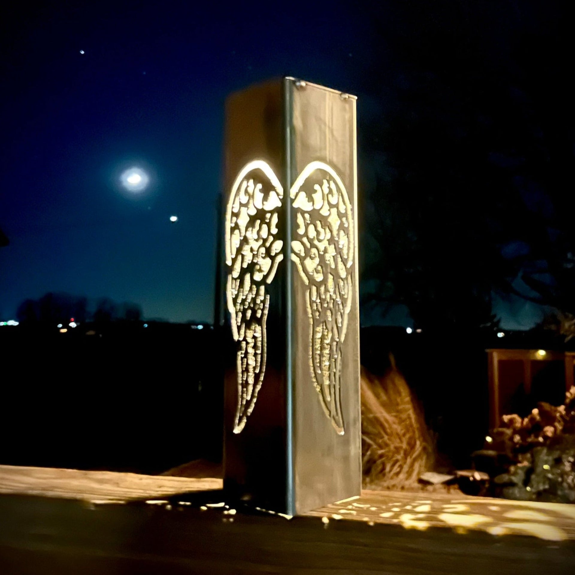 Decorative metal garden pillar with angel wings design under a night sky.