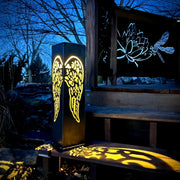 Decorative outdoor light pillar with angel wing design on a bench at night.