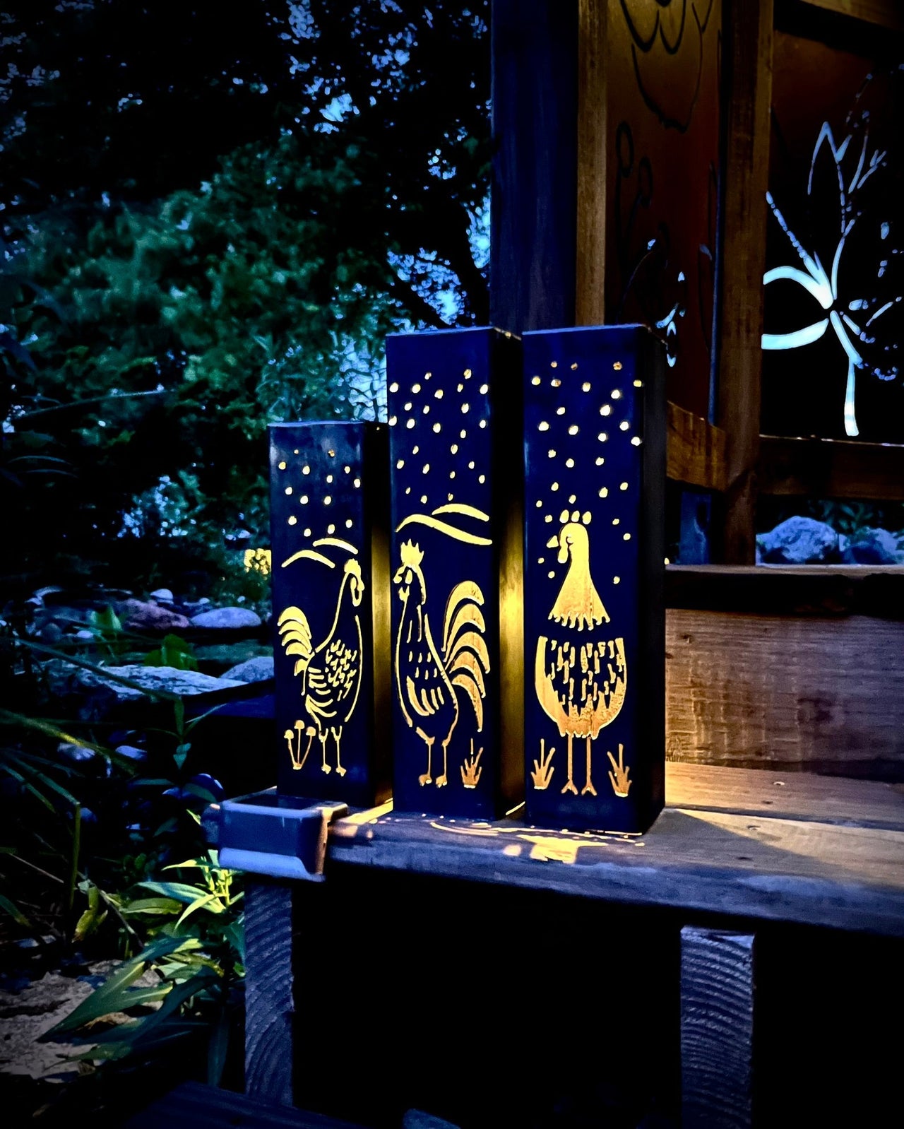 Three decorative Garden Column Pillars with rooster designs on a wooden surface outdoors.