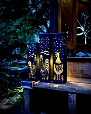 Three decorative Garden Column Pillars with rooster designs on a wooden surface outdoors.