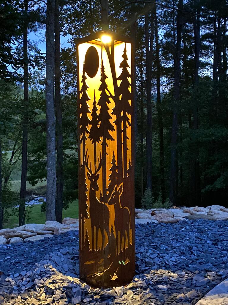 Decorative outdoor garden pillar with cut-out designs of trees and deer in a forest setting.
