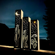 Three illuminated outdoor metal pillars with cactus and moon designs against a dark sky.