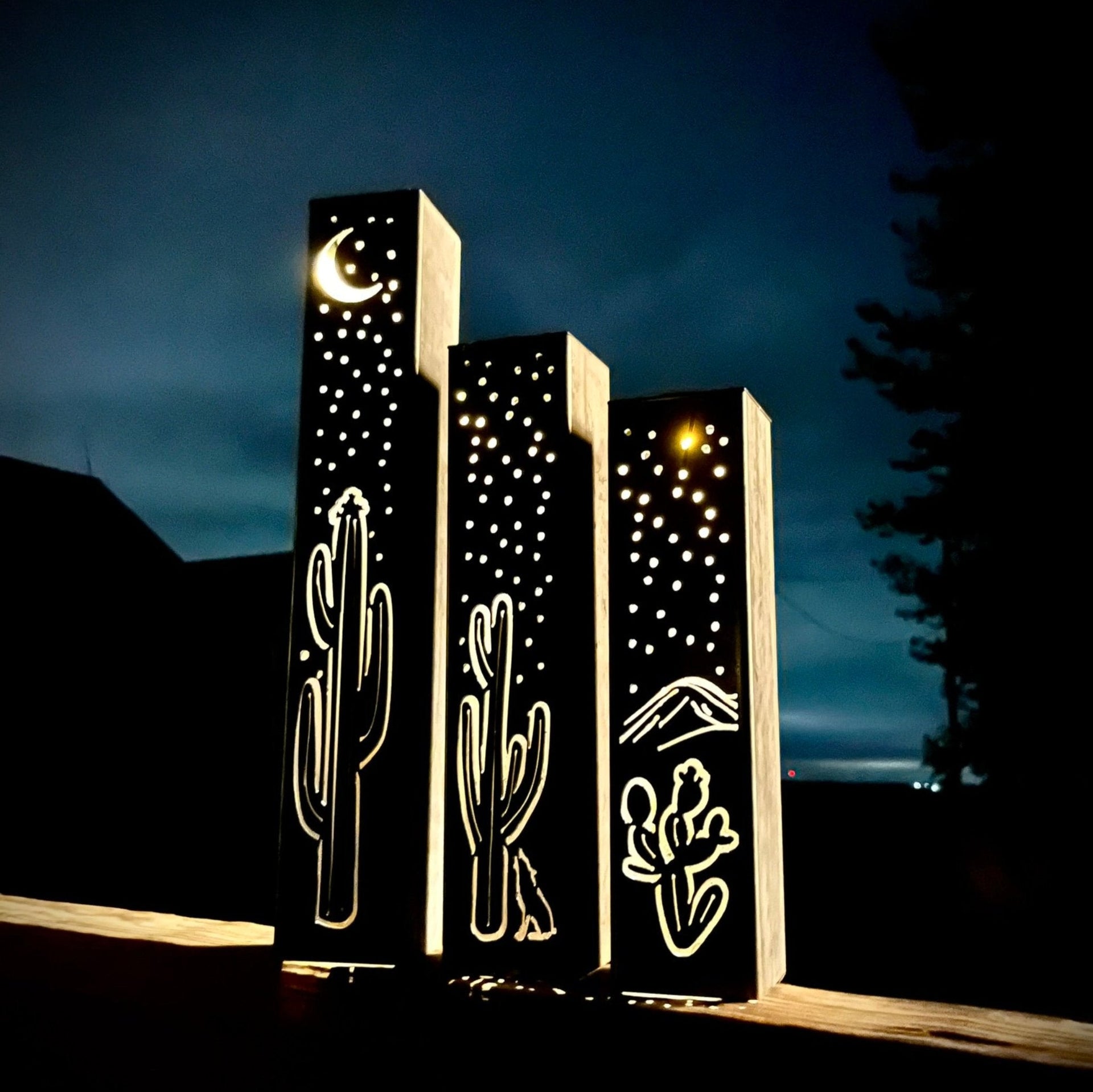 Three illuminated outdoor metal pillars with cactus and moon designs against a dark sky.