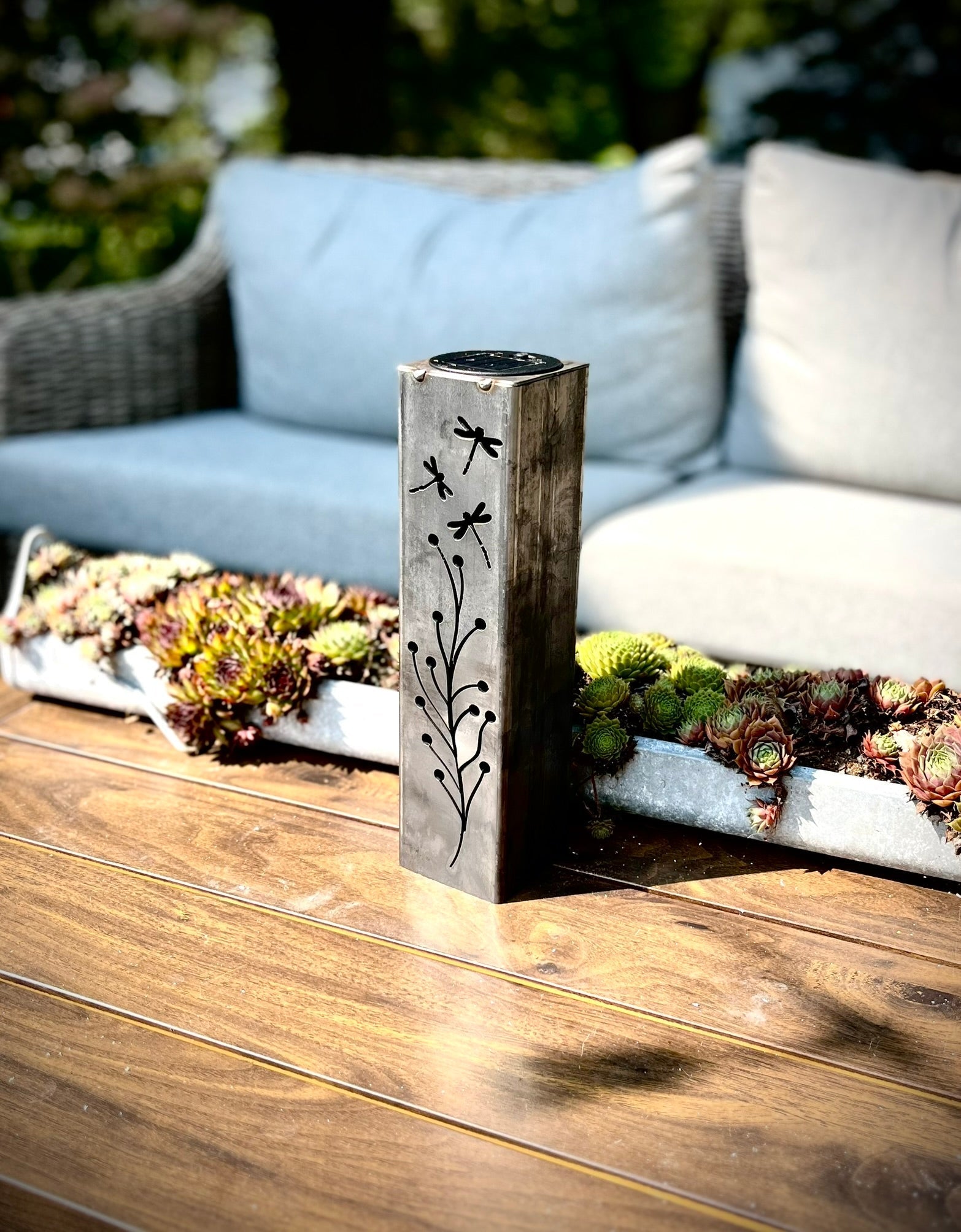 Decorative garden pillar with cut-out patterns on a wooden surface with a blurred outdoor background.