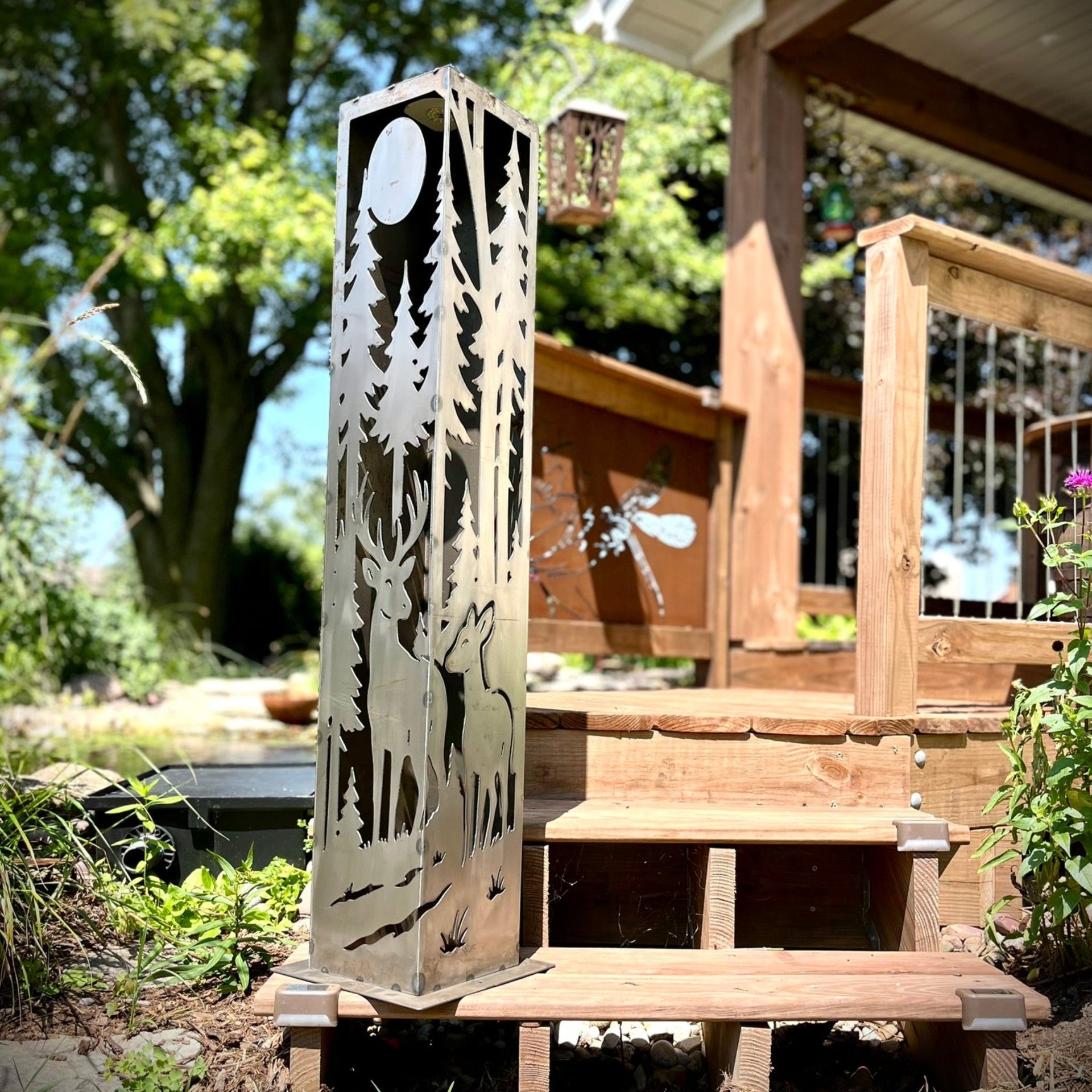 Decorative metal garden pillar with cut-out designs of deer and trees on a wooden deck.