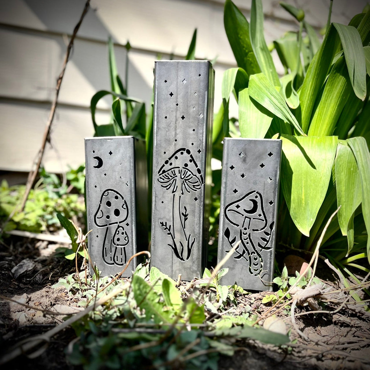 Three metal Garden Pillars with engraved designs of mushrooms and plants on a garden background.