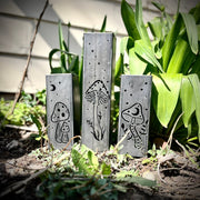Three metal Garden Pillars with engraved designs of mushrooms and plants on a garden background.