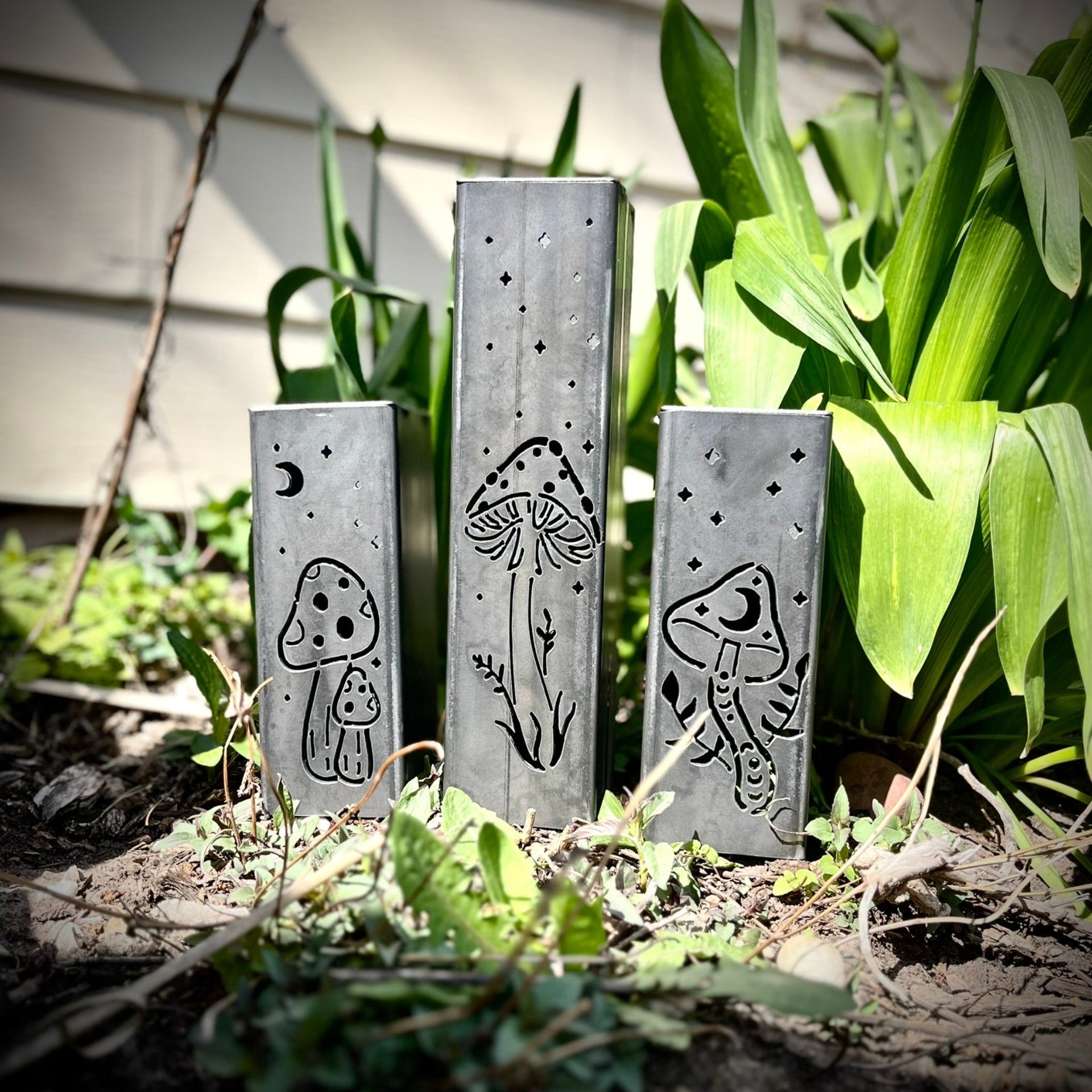 Three metal Garden Pillars with engraved designs of mushrooms and plants on a garden background.
