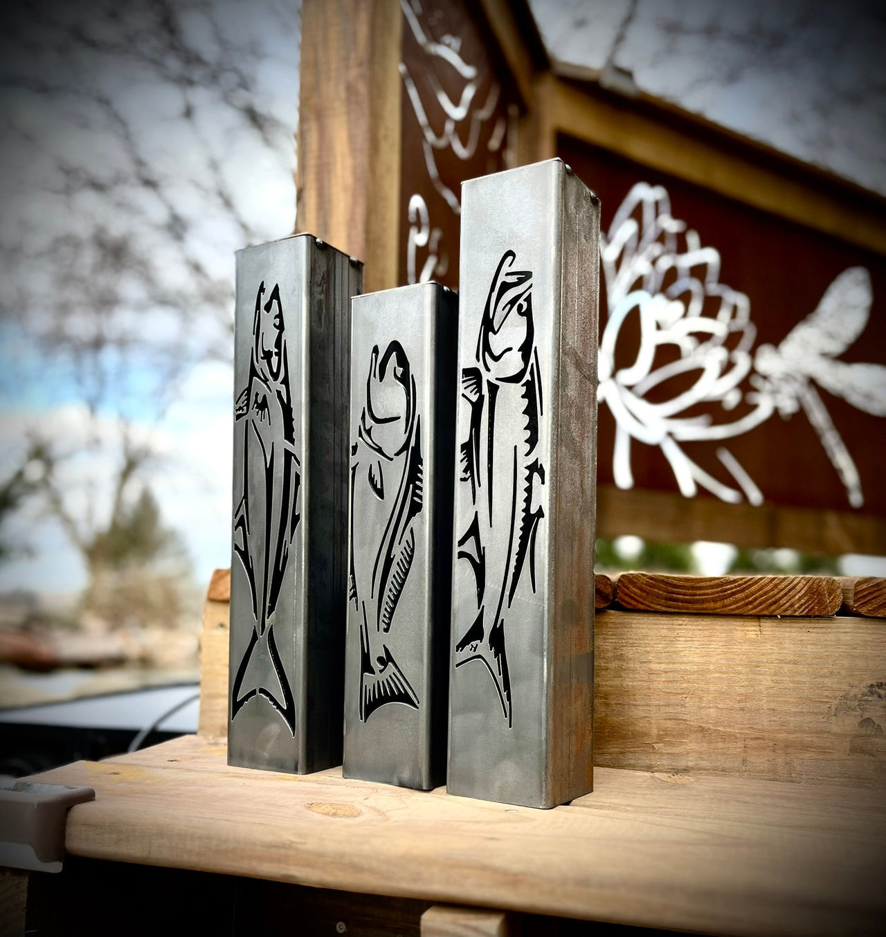 Three metal garden pillars  with fish designs on a wooden surface outdoors.