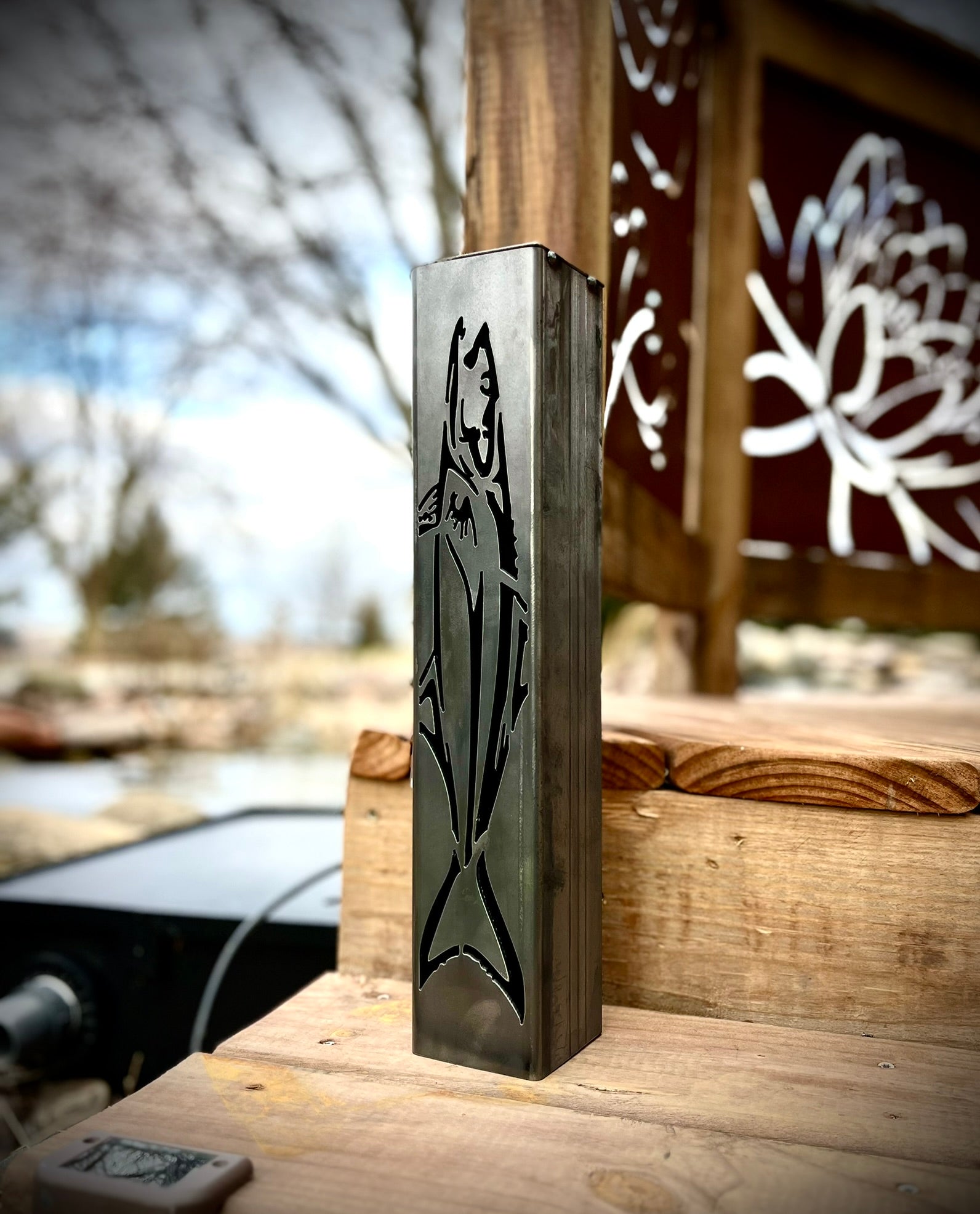 Metal garden pillar of a fish on a wooden surface with a blurred outdoor background