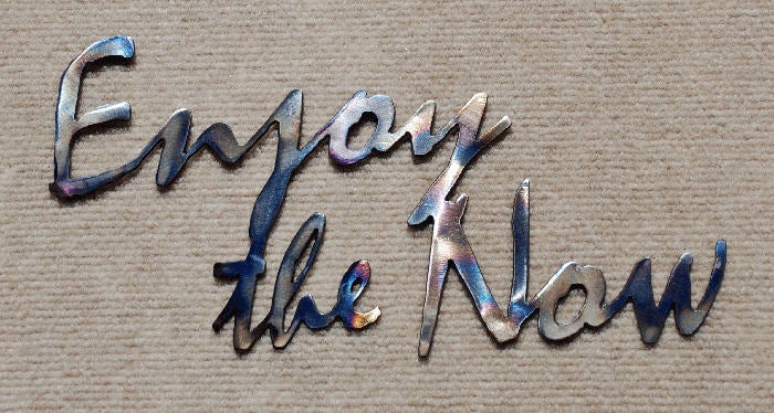 Decorative metal sign with 'Enjoy the Now' text on a textured beige background