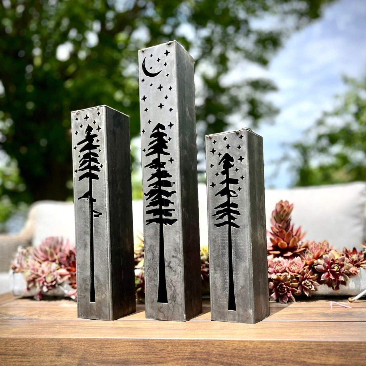 Three metal pillars with engraved tree designs on a wooden surface outdoors.
