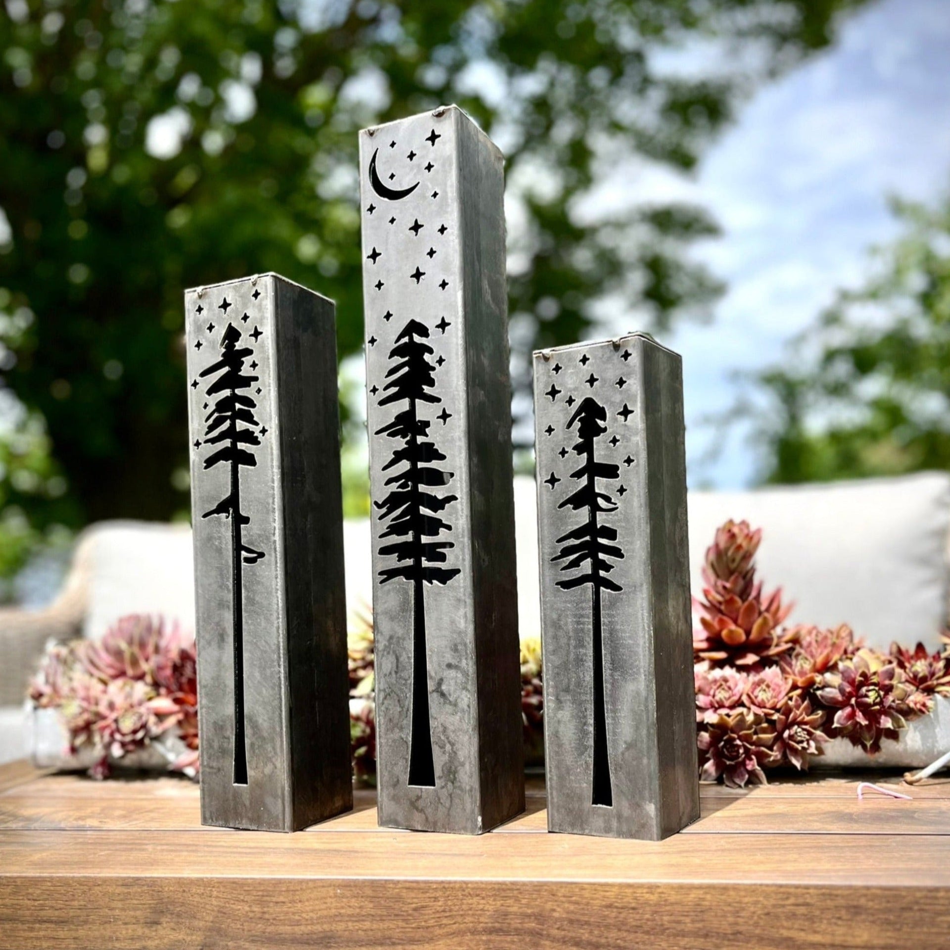 Three metal pillars with engraved tree designs on a wooden surface outdoors.