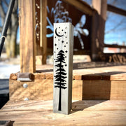Metal pillar with tree and moon design on a wooden surface outdoors.
