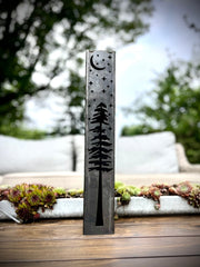Decorative metal pillars with tree and moon design on a wooden surface with blurred greenery in the background