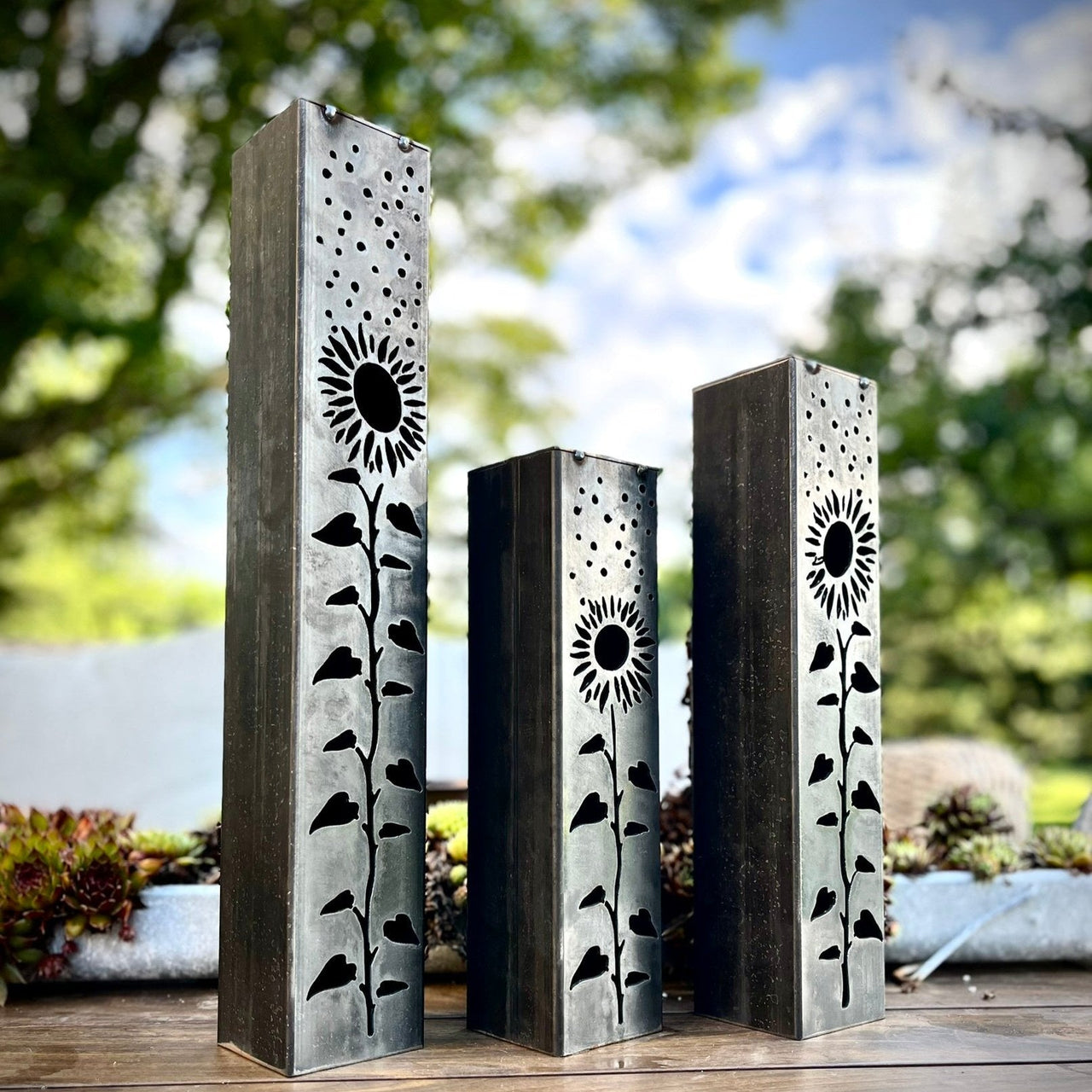 Three metal garden pillars with sunflower designs on a wooden surface outdoors.